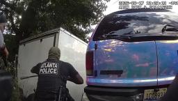 Atlanta Police officer crouching near a white trailer during an emergency search on Sylvan Road.