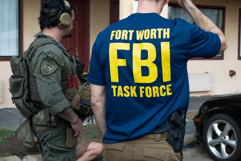 Rear view of an FBI task force member in a blue shirt with yellow lettering text reading Fort Worth FBI Task Force standing next to a tactical officer.
