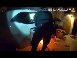 An APD officer reaches into a crashed white Dodge Challenger to perform a car fire rescue as flames burn on the ground below.