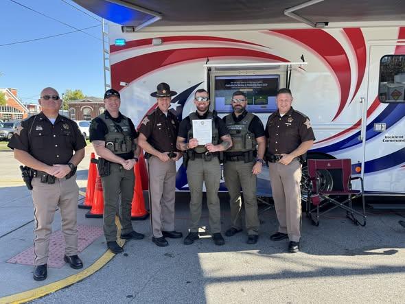 Six officers from the Floyd County Sheriff's Office stand proudly in front of a command vehicle, with Officer Anthony Whelan holding his Letter of Commendation.