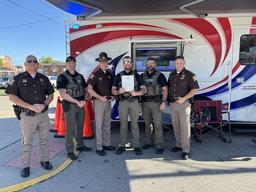 Six officers from the Floyd County Sheriff's Office stand proudly in front of a command vehicle, with Officer Anthony Whelan holding his Letter of Commendation.