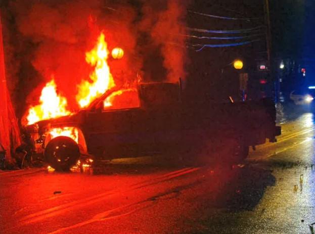 A black pickup truck engulfed in large orange flames after hitting a utility pole at night in Maynard.