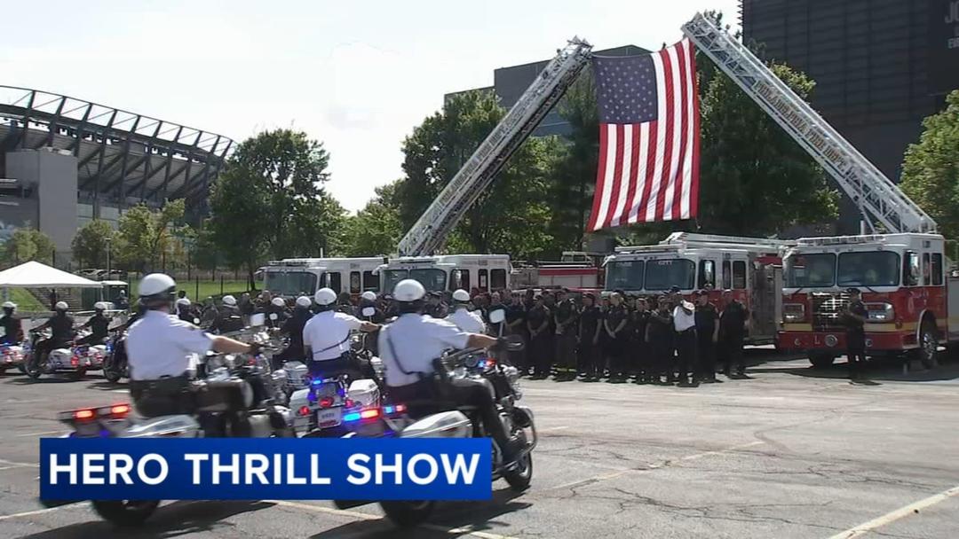 Two ladder trucks hold a large American flag above rows of firefighters at the Hero Thrill Show