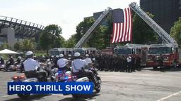 Two ladder trucks hold a large American flag above rows of firefighters at the Hero Thrill Show