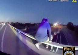 Police department bodycam screenshot depicting an officer approaching a person positioned on a highway overpass railing.