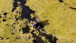 A person in a dark shirt with a blurred face is in the water of a pond, seen from above.