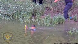 A Fort Worth police officer approaches a child holding a yellow object while partially submerged in water near shoreline weeds.