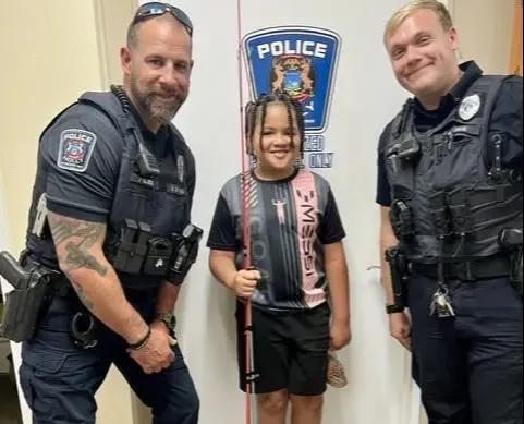 Two Holly police officers stand in the department lobby beside a young boy holding new fishing rods.