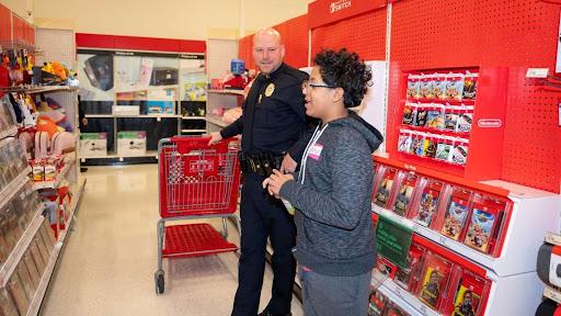 A Raleigh police officer smiling while helping a young boy shop with a red cart at Target.