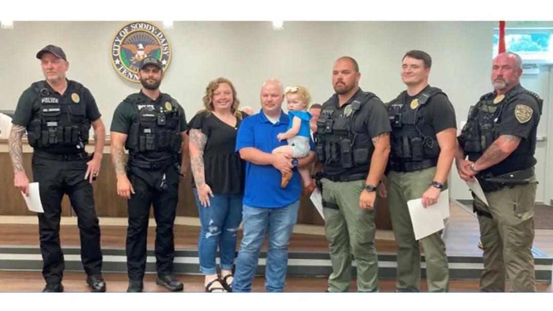 Five Soddy Daisy police officers stand with a mother and father holding a toddler girl inside a city building.